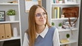 A young, professional woman with blonde hair and glasses smiles subtly in a modern office setting, exuding calm and confidence