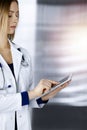 Young professional woman-doctor is using a tablet computer, while standing in a sunny clinic. Portrait of beautiful Royalty Free Stock Photo