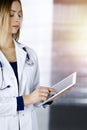 Young professional woman-doctor is using a tablet computer, while standing in a sunny clinic. Portrait of beautiful Royalty Free Stock Photo