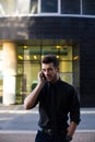 Young proud businessman having smartphone conversation while standing outside his corporation. Royalty Free Stock Photo