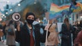 Young rebellious woman with face mask and megaphone holding rainbow flag in crowd. Protest against discrimination