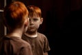 A young red-haired boy looks thoughtfully at himself in the mirror on a dark background. Childhood crisis, depression concept. Royalty Free Stock Photo