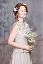Young red-haired bride in pearl-gray dress. She stands, her eyes are dreamy closed, she is holding a bouquet of white wildflowers.