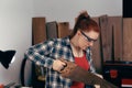 Young red-haired woman, carpenter, sawing a wooden board with a saw, in her small carpentry workshop.