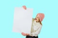 A young red-haired girl in a knitted pink hat and mittens holds a large white poster with both hands and looks at the Royalty Free Stock Photo