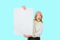 A young red-haired girl in a knitted pink hat and mittens holds a large white poster with both hands and looks to the Royalty Free Stock Photo