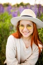 Young red haired girl with long hair in a wicker hat with a ribbon. Woman sitting with on a field of lupins at sunset and smiling