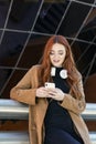 young red-haired girl looking at her cell phone, with wireless headphones leaning on a railing on a city street Royalty Free Stock Photo