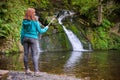 Young woman blogger in autumn mountains near lake and waterfall is recording video, taking photo with selfie stick Royalty Free Stock Photo