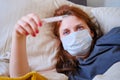 Young red-haired woman looks at a thermometer while lying in bed. Sick girl lies at home in a medical mask Royalty Free Stock Photo