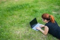 Young red-haired woman remotely works out lying on green grass. Freelancer girl prints on a laptop outdoors.