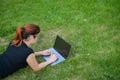 Young red-haired woman remotely works out lying on green grass. Freelancer girl prints on a laptop outdoors.