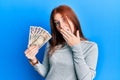 Young red head girl holding japanese yen banknotes covering mouth with hand, shocked and afraid for mistake