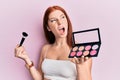 Young red head girl holding makeup brush and blush palette angry and mad screaming frustrated and furious, shouting with anger Royalty Free Stock Photo