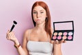 Young red head girl holding makeup brush and blush palette puffing cheeks with funny face