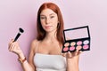 Young red head girl holding makeup brush and blush palette relaxed with serious expression on face Royalty Free Stock Photo