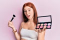 Young red head girl holding makeup brush and blush palette in shock face, looking skeptical and sarcastic, surprised with open Royalty Free Stock Photo