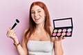 Young red head girl holding makeup brush and blush palette smiling and laughing hard out loud because funny crazy joke Royalty Free Stock Photo