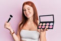 Young red head girl holding makeup brush and blush palette winking looking at the camera with expression, cheerful and happy Royalty Free Stock Photo