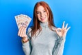 Young red head girl holding 500 mexican pesos banknotes doing ok sign with fingers, smiling friendly gesturing excellent symbol