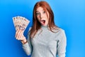 Young red head girl holding 500 mexican pesos banknotes scared and amazed with open mouth for surprise, disbelief face