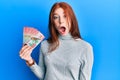 Young red head girl holding 100 new zealand dollars banknote scared and amazed with open mouth for surprise, disbelief face Royalty Free Stock Photo