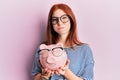 Young red head girl holding piggy bank with glasses puffing cheeks with funny face