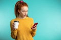 Young redhaired woman with coffee and smartphone isolated over b