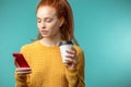 Young redhaired woman with coffee and smartphone isolated over blue background