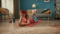 A young redhaired woman in pink sportswear lies on her stomach on the floor in the living room. A woman holds a phone in