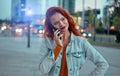 Young redhaired woman standing at night with phone by european skyline