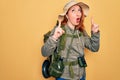 Young redhead backpacker woman hiking wearing backpack and hat over yellow background amazed and surprised looking up and pointing Royalty Free Stock Photo