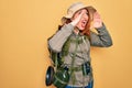 Young redhead backpacker woman hiking wearing backpack and hat over yellow background Shouting angry out loud with hands over