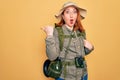 Young redhead backpacker woman hiking wearing backpack and hat over yellow background Surprised pointing with hand finger to the Royalty Free Stock Photo