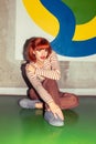 Young redhead Caucasian woman in retro style of 70s sitting at wall vertical looking into camera Royalty Free Stock Photo