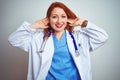 Young redhead doctor woman using stethoscope over white isolated background Smiling pulling ears with fingers, funny gesture