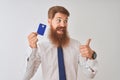 Young redhead irish businessman holding credit card standing over isolated white background pointing and showing with thumb up to Royalty Free Stock Photo