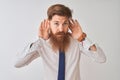 Young redhead irish businessman standing over isolated white background Trying to hear both hands on ear gesture, curious for Royalty Free Stock Photo