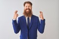 Young redhead irish businessman wearing suit standing over isolated white background gesturing finger crossed smiling with hope