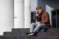 Young redhead man in brown jacket is talking by phone while sitting on steps outdoors. Royalty Free Stock Photo