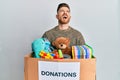 Young redhead man holding donation box with toys angry and mad screaming frustrated and furious, shouting with anger looking up Royalty Free Stock Photo