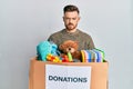 Young redhead man holding donation box with toys puffing cheeks with funny face