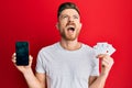 Young redhead man holding smartphone and casino card angry and mad screaming frustrated and furious, shouting with anger looking Royalty Free Stock Photo