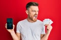 Young redhead man holding smartphone and casino card angry and mad screaming frustrated and furious, shouting with anger Royalty Free Stock Photo