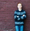 Young redhead man in a sweater and jeans standing next to a brick wall with folded arms