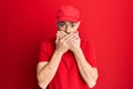 Young redhead man wearing delivery uniform and cap shocked covering mouth with hands for mistake