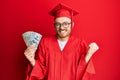 Young redhead man wearing graduation robe holding dollars screaming proud, celebrating victory and success very excited with Royalty Free Stock Photo