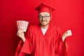 Young redhead man wearing graduation robe holding 20 shekels screaming proud, celebrating victory and success very excited with Royalty Free Stock Photo