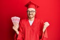 Young redhead man wearing graduation robe holding 100 yuan screaming proud, celebrating victory and success very excited with Royalty Free Stock Photo