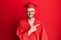 Young redhead man wearing red graduation cap and ceremony robe cheerful with a smile of face pointing with hand and finger up to Royalty Free Stock Photo